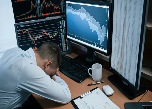 A trader rests his forehead on his desk surrounded by market monitors showing steep declines.